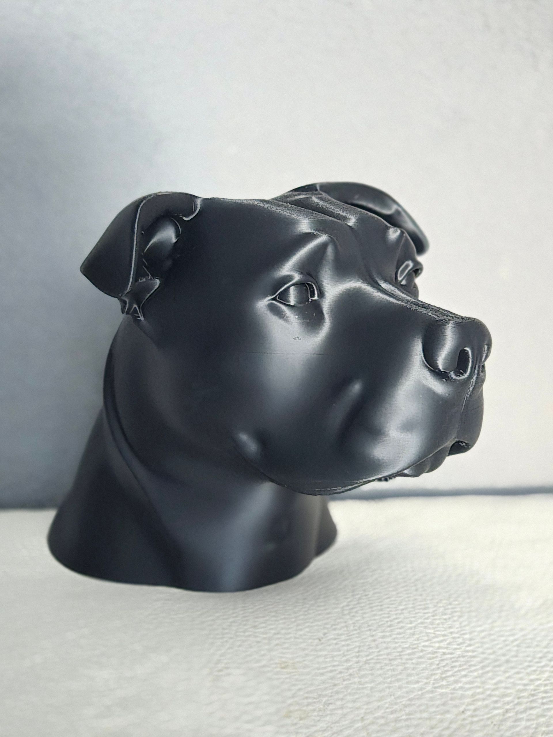 Een gladde, zwarte sculptuur met matte afwerking van de kop van een Staffordshire Bull Terrier tegen een grijs-witte achtergrond.