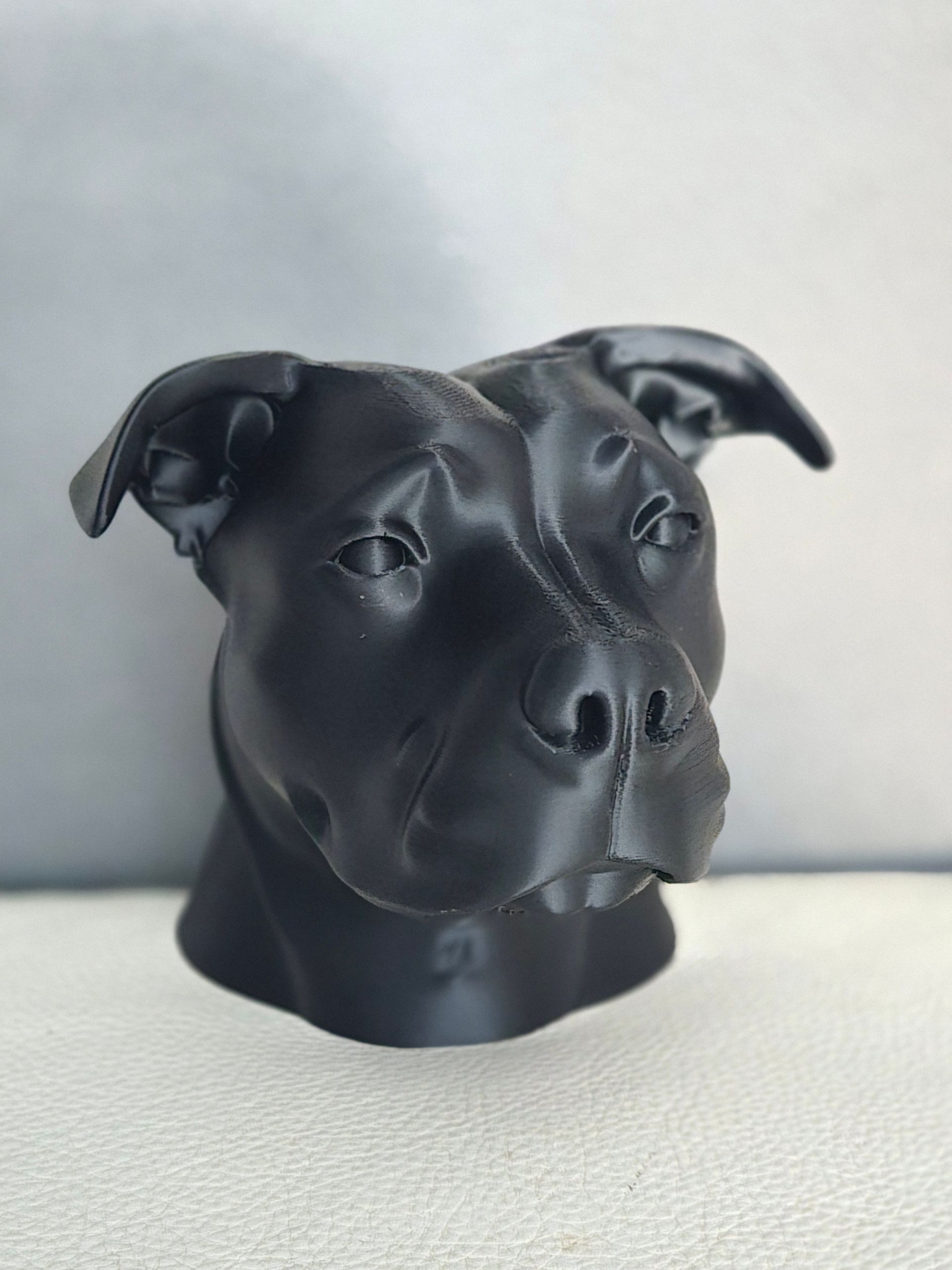 Een zwart, gebeeldhouwd buste van een pitbullkop, die iets naar rechts kijkt, tegen een effen, lichte achtergrond.
