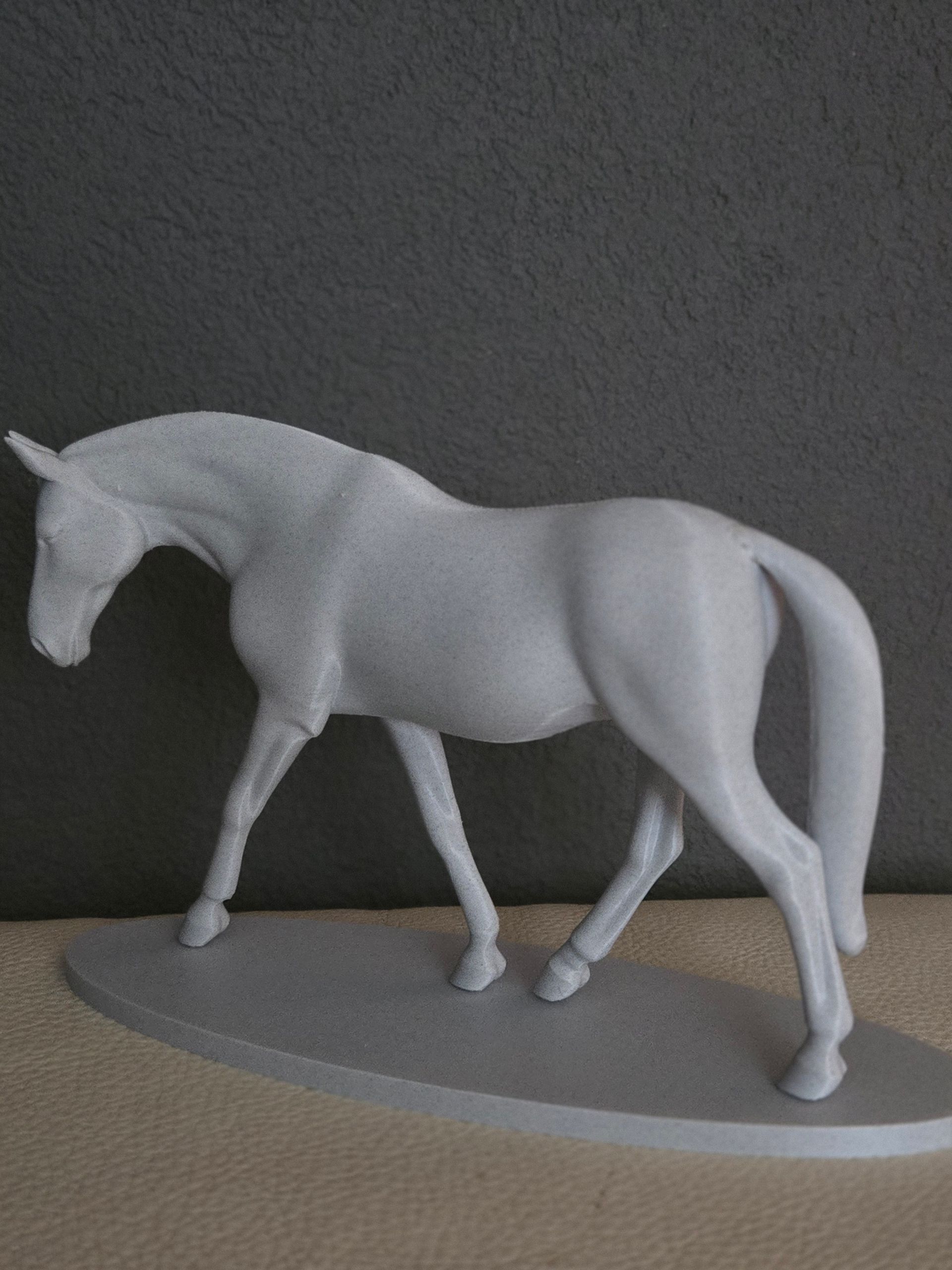 Een grijs, ongeverfd modelpaardje staat op een ovale sokkel tegen een donkergrijze achtergrond.