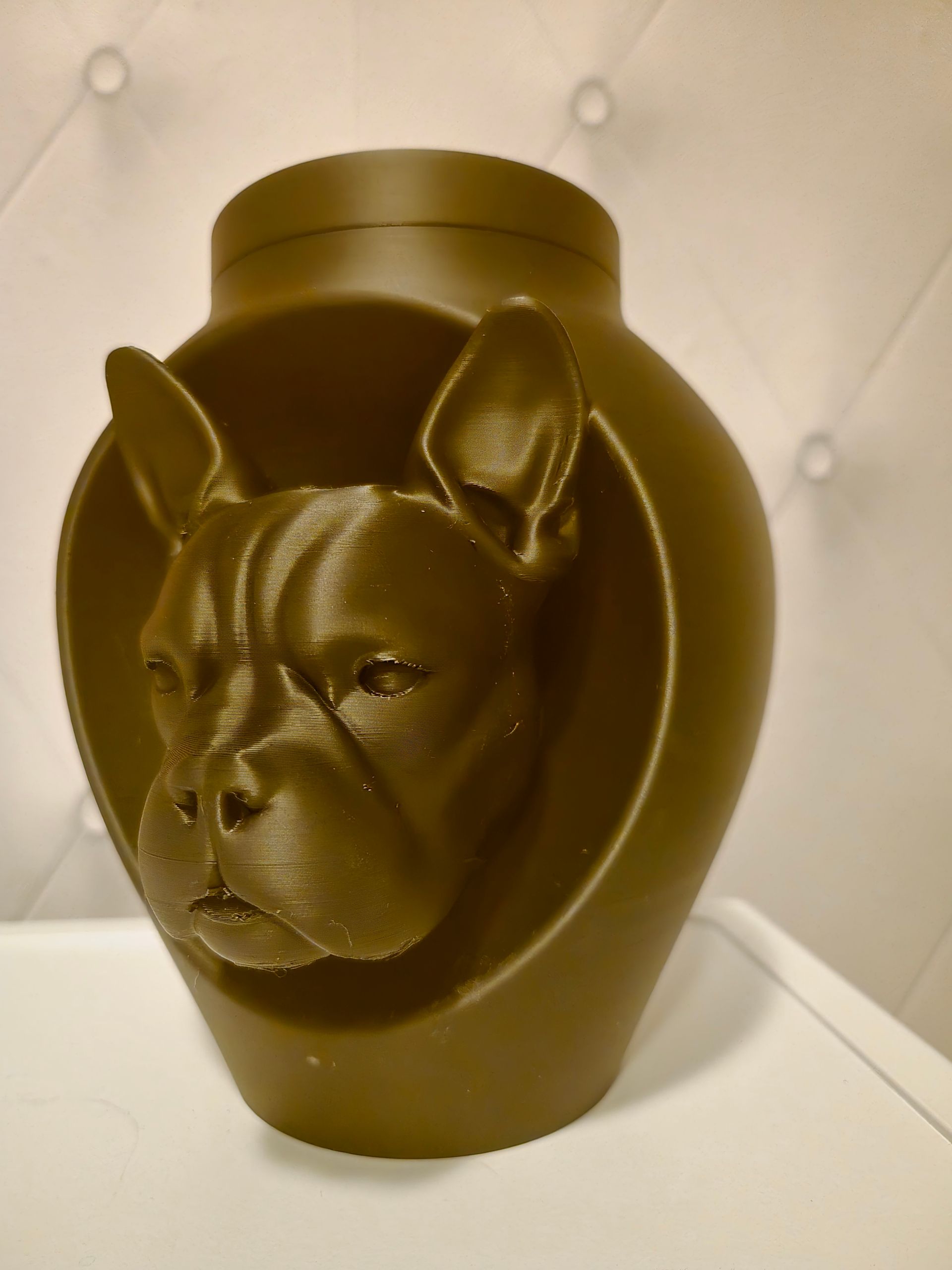Een donkerbruine, matte urn of vaas met een 3D-gevormde kop van een Franse bulldog in het midden van de voorkant, geplaatst tegen een gecapitonneerde muur.