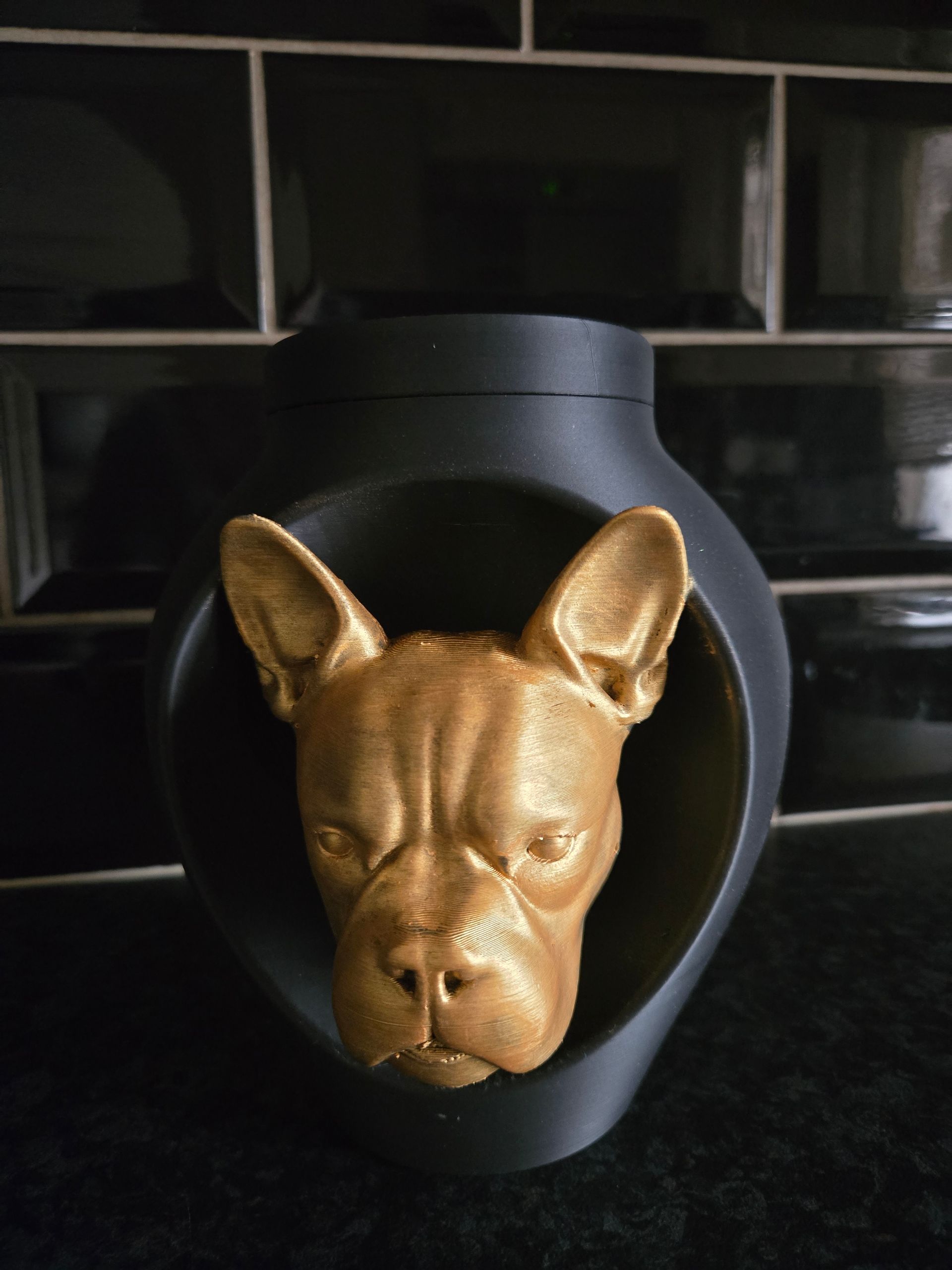 Een matzwarte keramische pot met een gebeeldhouwde, goudgeverfde kop van een Franse bulldog in het midden van een ronde uitsparing.