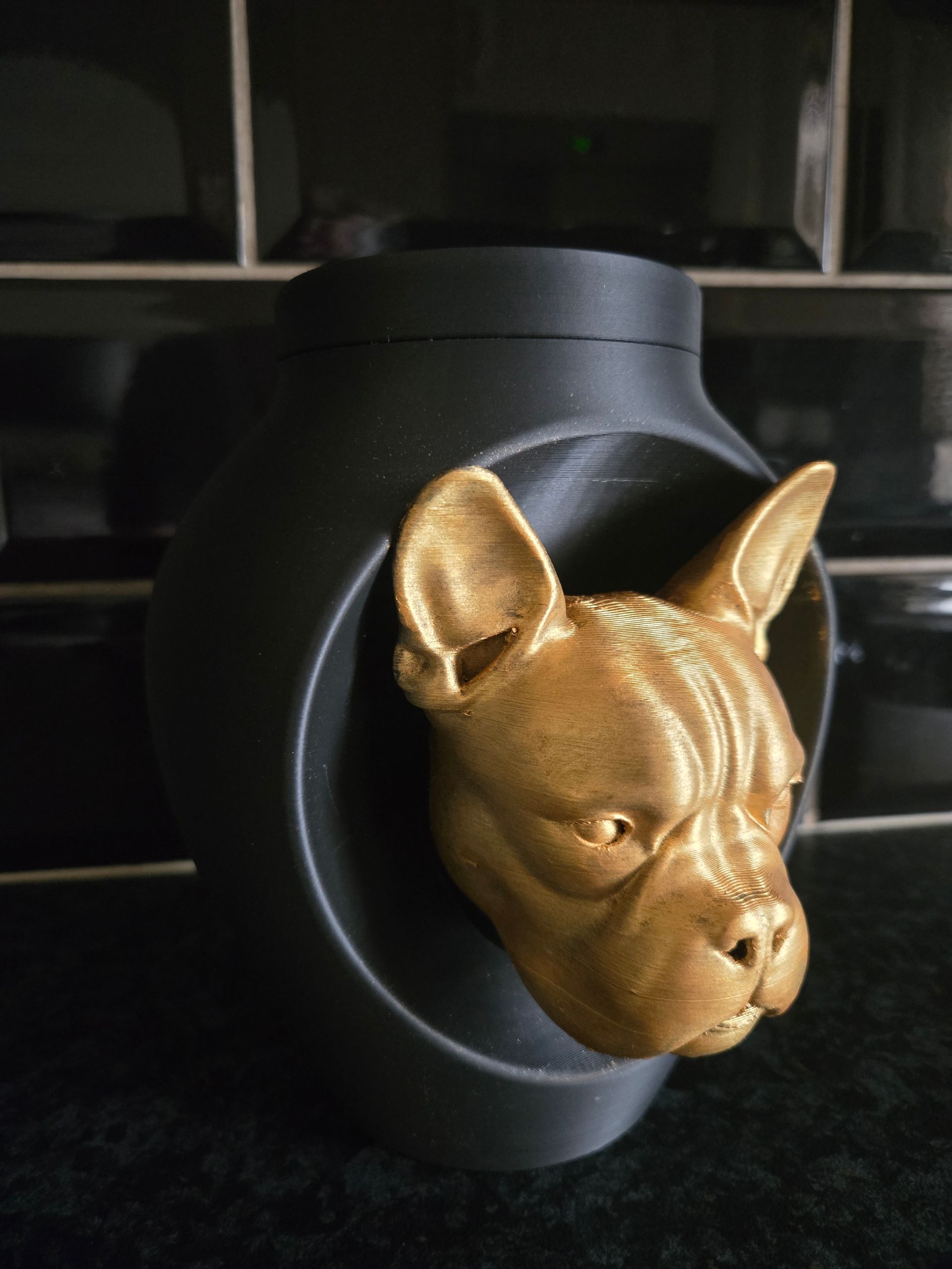 Een matzwarte vaas heeft een goudkleurige, 3D-geprinte bulldogkop die uit een ronde uitsparing aan de voorkant steekt.
