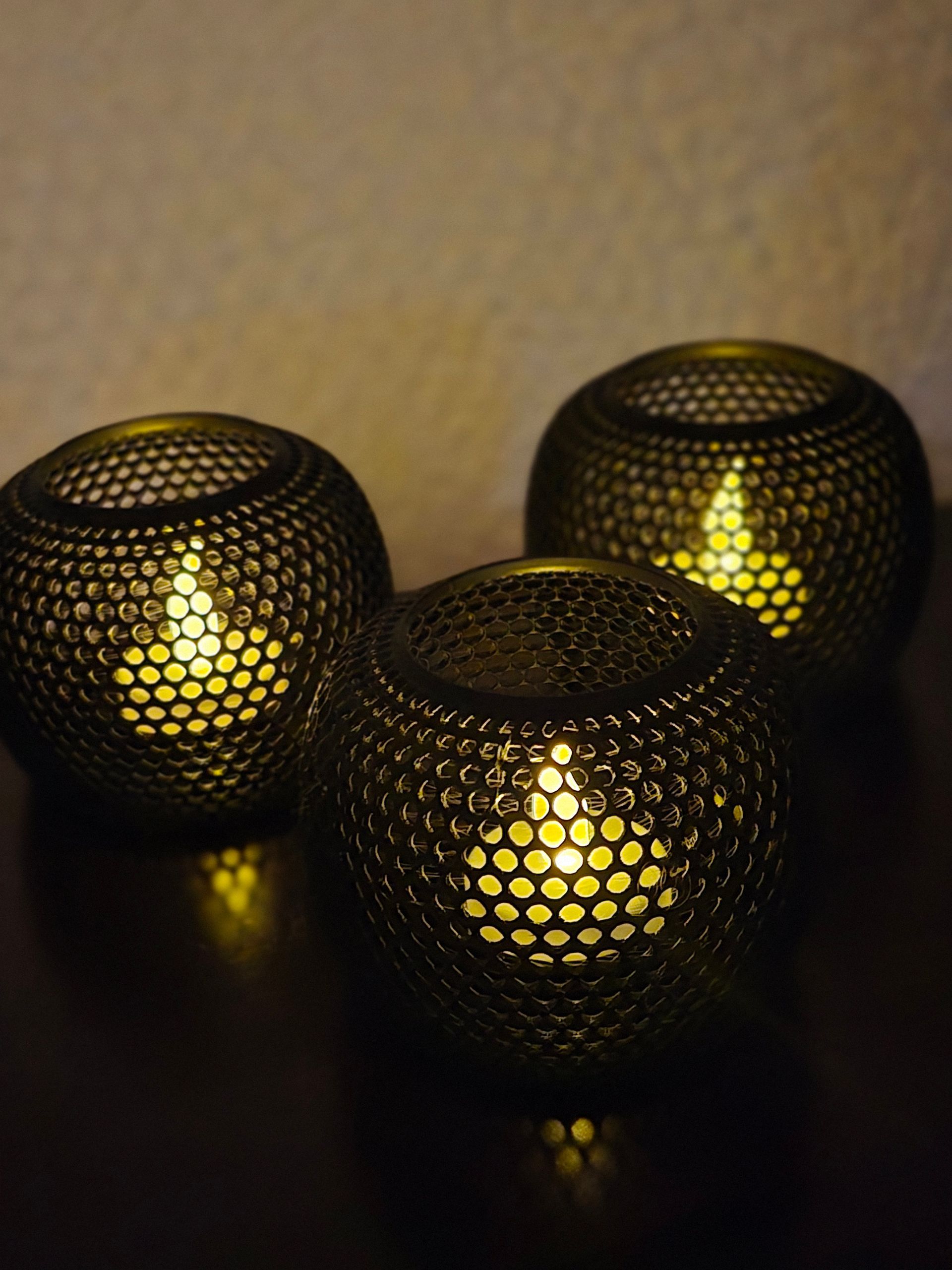 Drie donkere, metalen, bolvormige kaarsenhouders met een geperforeerd honingraatpatroon, die een warm licht verspreiden op een donkere ondergrond.