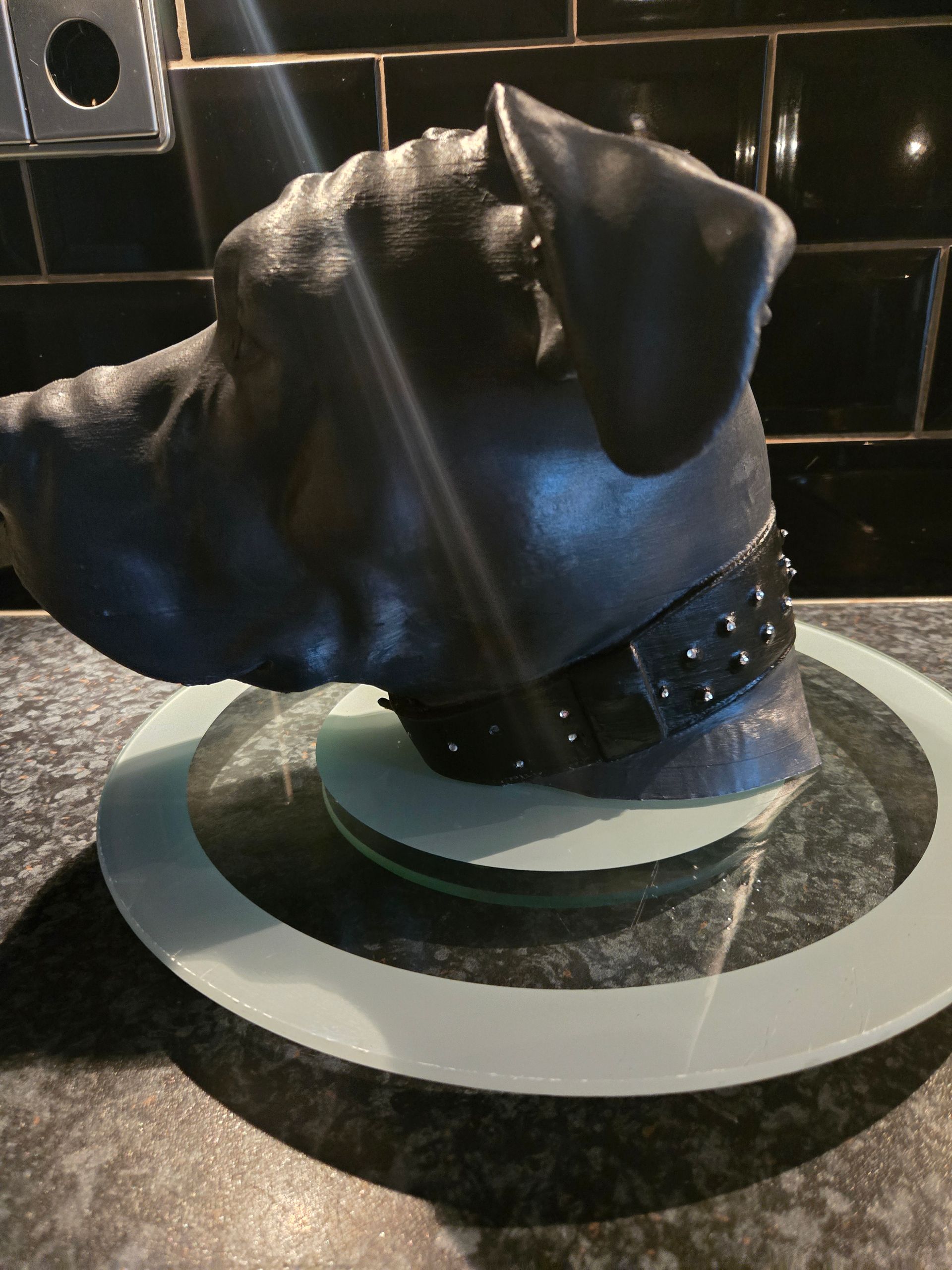 Een matzwarte sculptuur van een hondenkop met een met studs bezette leren halsband, staand op een glazen sokkel tegen een betegelde achterwand.
