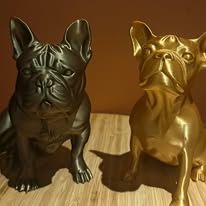 Twee gestileerde Franse bulldogbeeldjes, een donkergrijs en een goudkleurig, staan ​​naast elkaar op een licht houten oppervlak.