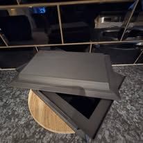 Een matzwarte, rechthoekige haard- of kacheldeur met frame, rustend op een houten oppervlak met structuur.