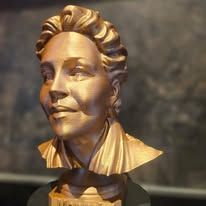 Een bronzen buste van een persoon met golvend haar, die iets naar links kijkt, tegen een donkere, wazige achtergrond.