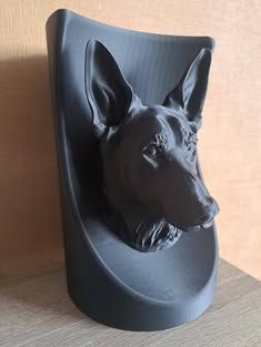 Een zwart, 3D-geprint hoofd van een Duitse herder, gemonteerd op een gebogen, bijpassende sokkel tegen een licht houten achtergrond.