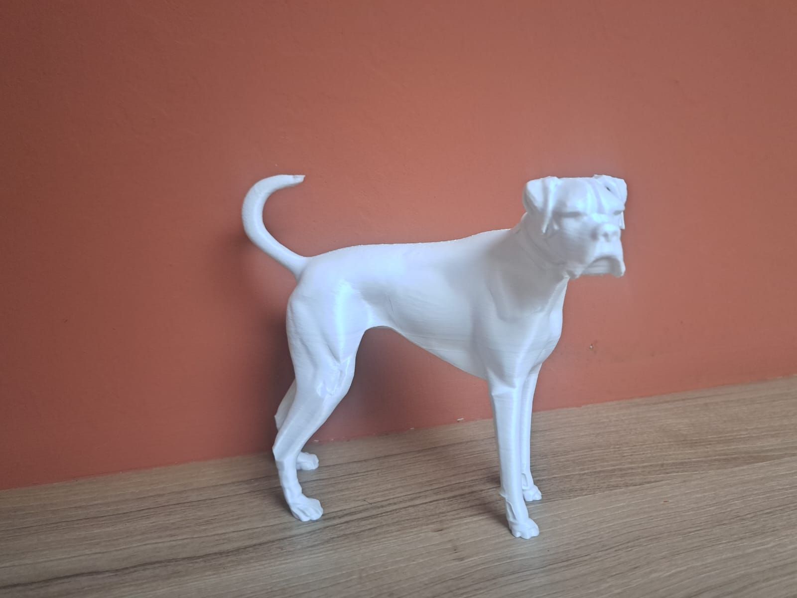 Een wit, 3D-geprint beeld van een boxerhond, staand op een houten ondergrond tegen een terracotta-kleurige muur.