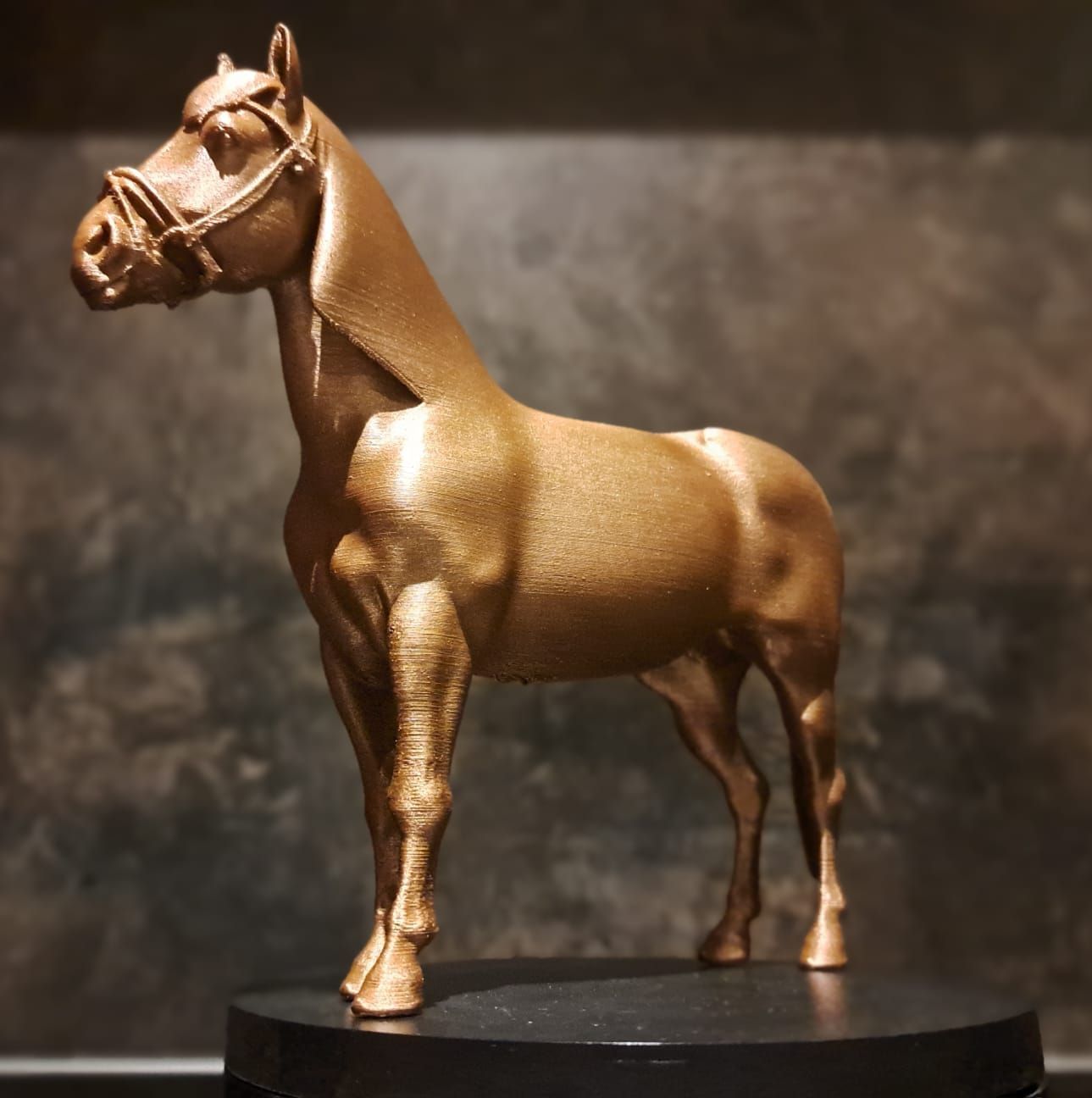 Een beeldje van een paard met een hoofdstel, uitgevoerd in metaalgoud, staat op een ronde zwarte sokkel tegen een donkere, gestructureerde achtergrond.
