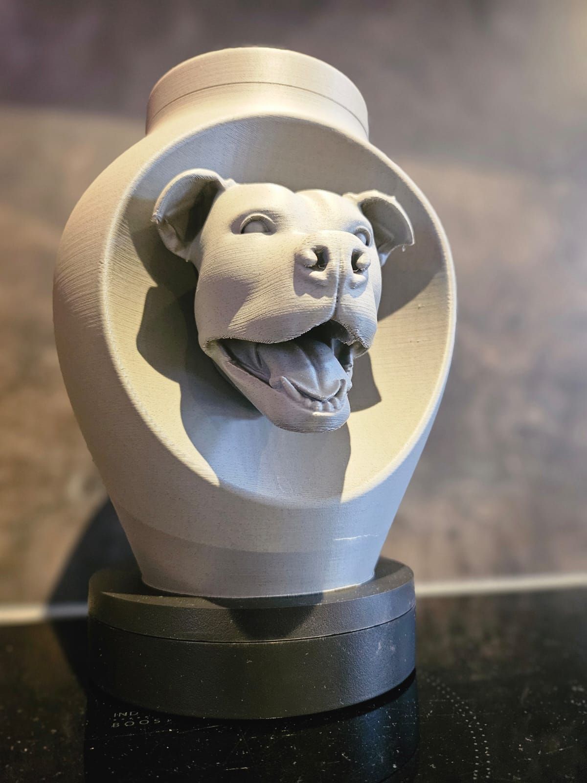 Een 3D-geprinte lichtgrijze urn met een gebeeldhouwde hondenkop in een cirkelvormig frame, geplaatst op een donkere, cilindrische voet.