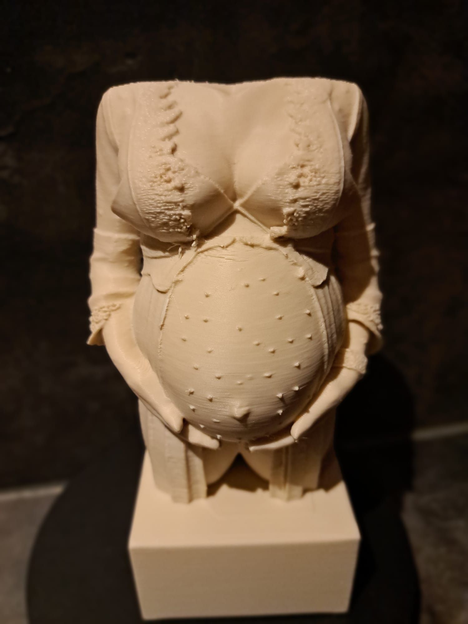 Een lichtgekleurd torso-beeld van een zwangere vrouw met een kanten topje aan, haar handen om haar buik, op een vierkante sokkel.