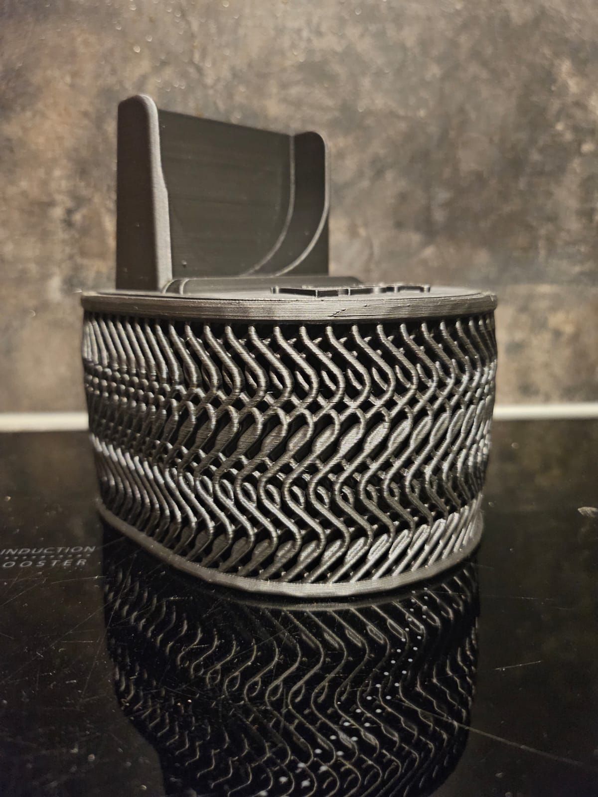 Een 3D-geprint object van zwart plastic met een complexe, rasterachtige ondersteuningsstructuur, rustend op een donker, reflecterend oppervlak.