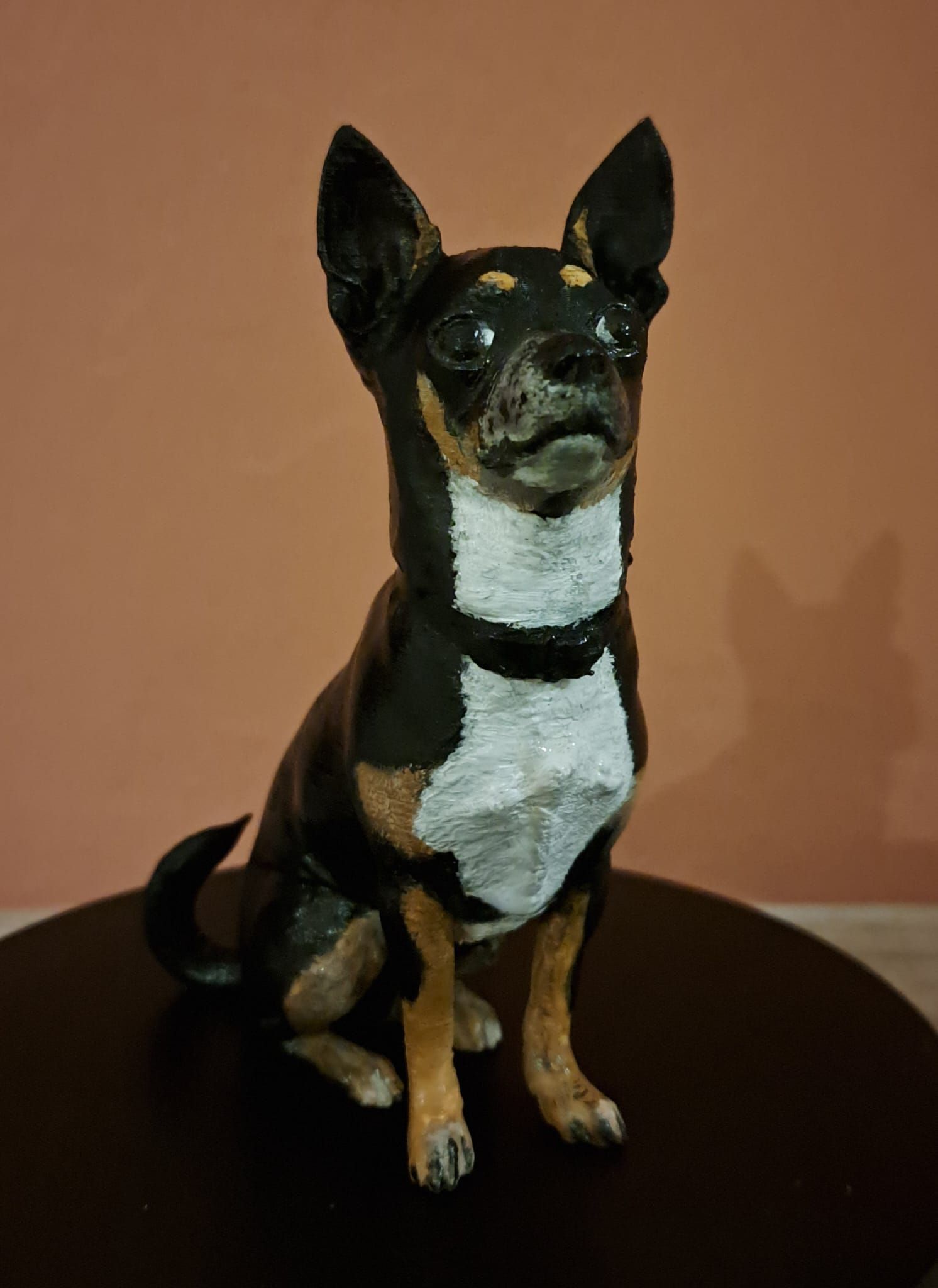 Een beeldje van een zwart-bruine chihuahua met een witte borst en halsband, zittend op een donkere ondergrond tegen een perzikkleurige muur.