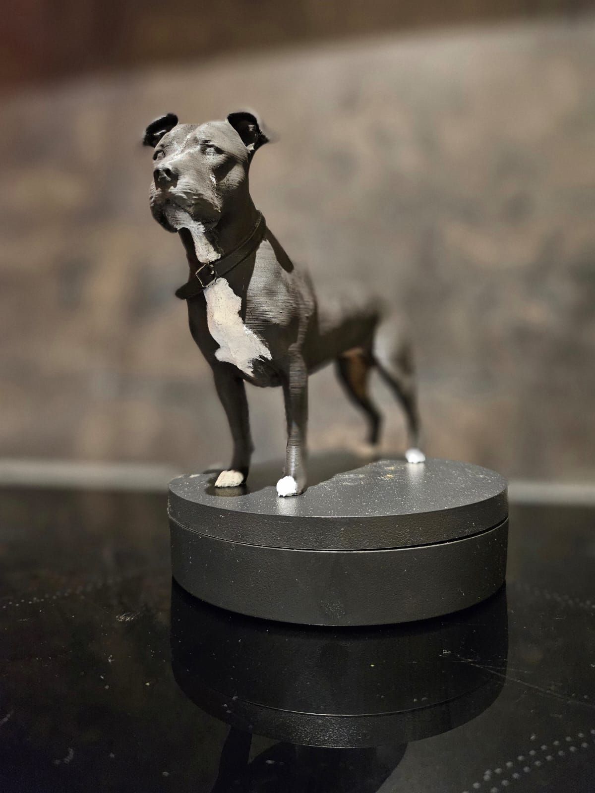 Een gedetailleerd beeldje van een grijze hond met een witte vlek op zijn borst staat op een ronde, donkergrijze sokkel tegen een wazige muur.