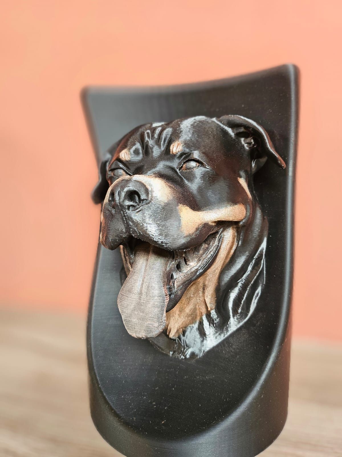 Een realistische 3D-sculptuur van een Rottweilerkop met uitgestoken tong, gemonteerd op een zwarte, gebogen plaat tegen een oranje muur.