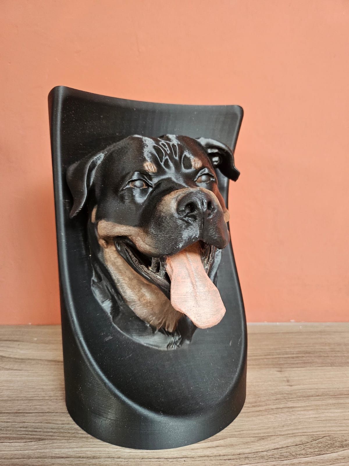 Een zwarte, 3D-geprinte gebogen muurbeugel met een realistische Rottweilerkop inclusief uitgestoken tong.