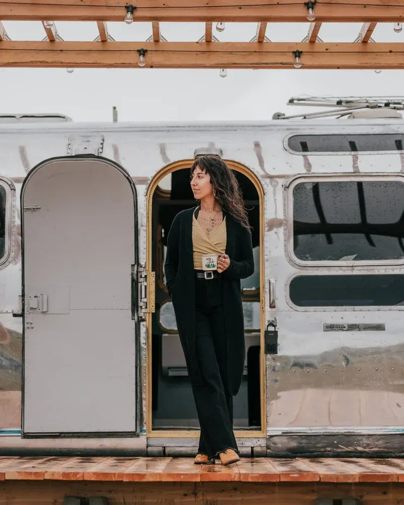 a woman is standing in front of an airstream holding a cup of coffee .