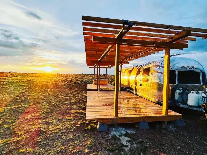 an airstream trailer is parked under a wooden pergola in the middle of a field .