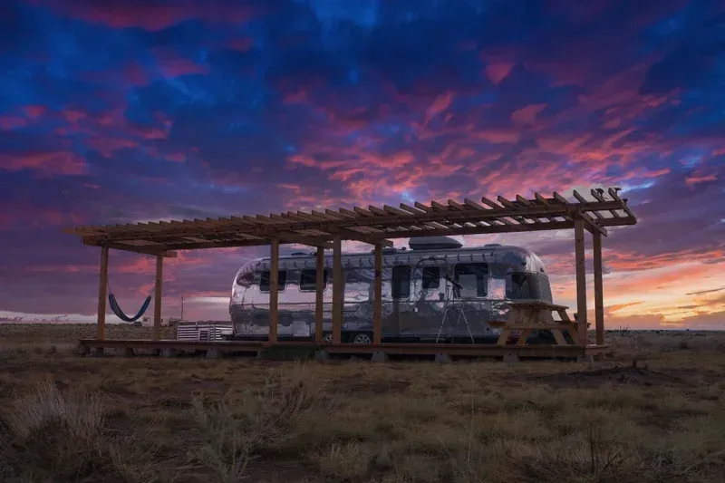 an airstream is parked under a pergola in the middle of a field at sunset .