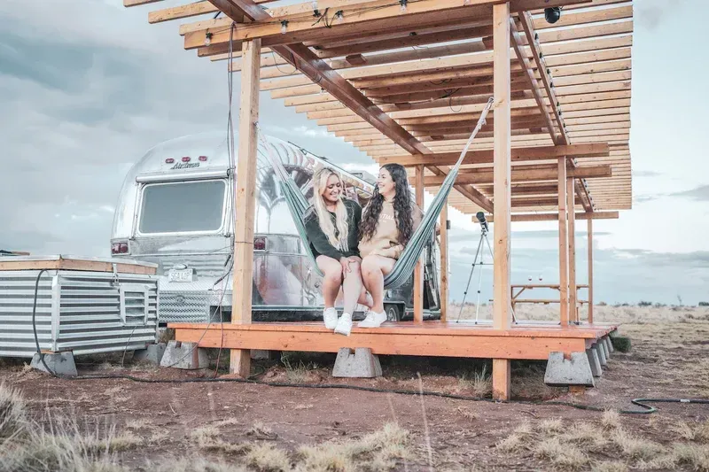 two women are sitting in a hammock on a porch next to an airstream .