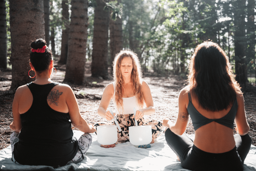 a certified sound healer plays sound bowls for two participants sitting cross-legged on a picnic blanket in the forest