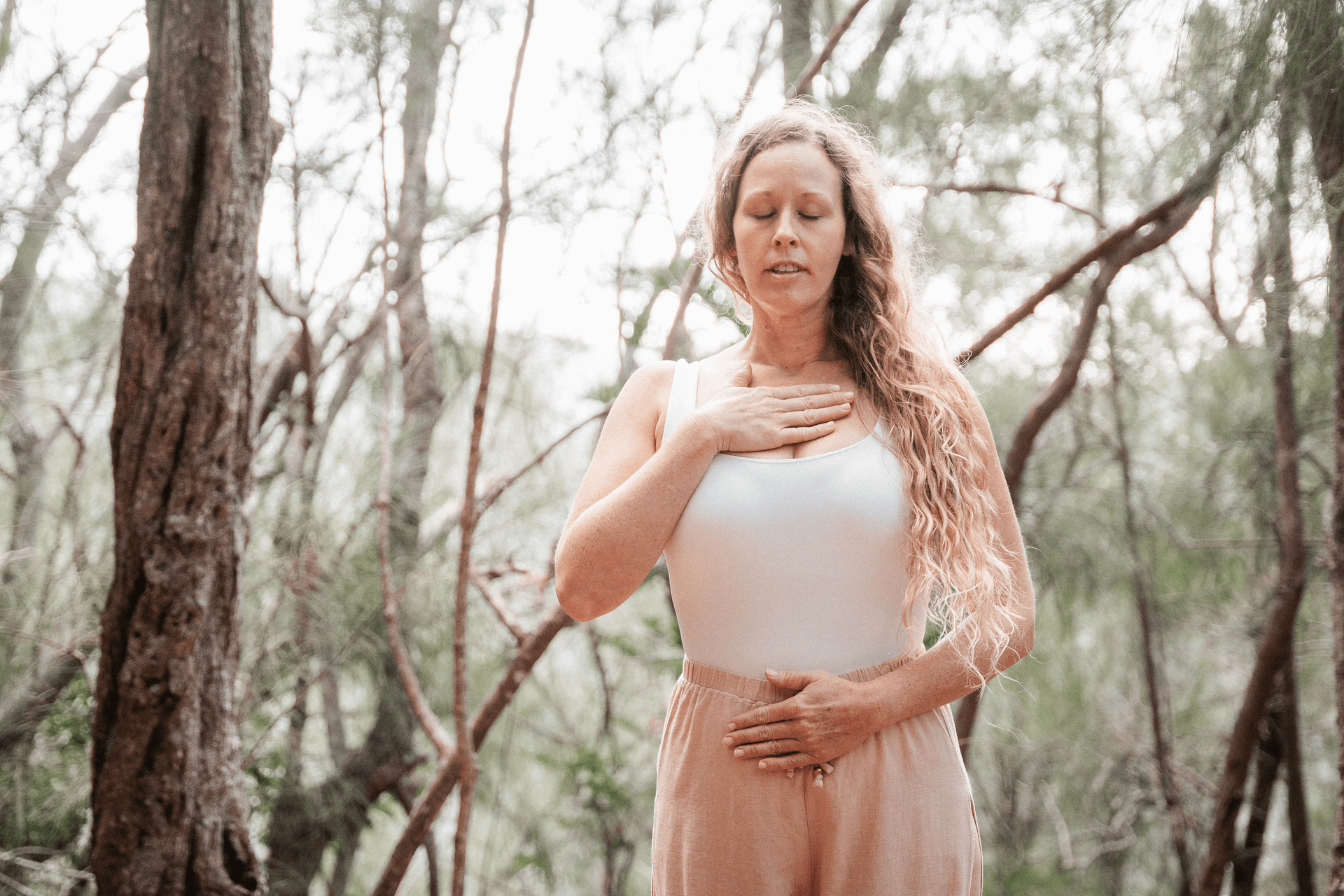 sound healing practitioner stands in the forest with one hand over her heart and the other over her belly