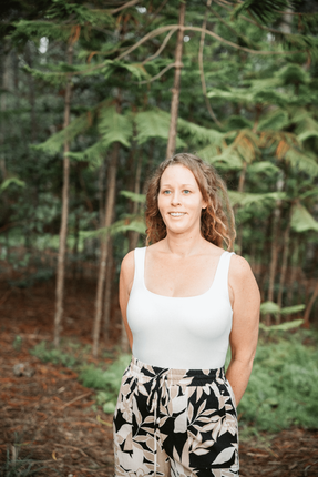 reiki master jessica standing in a green forest wearing a white tank top smiling in the distance