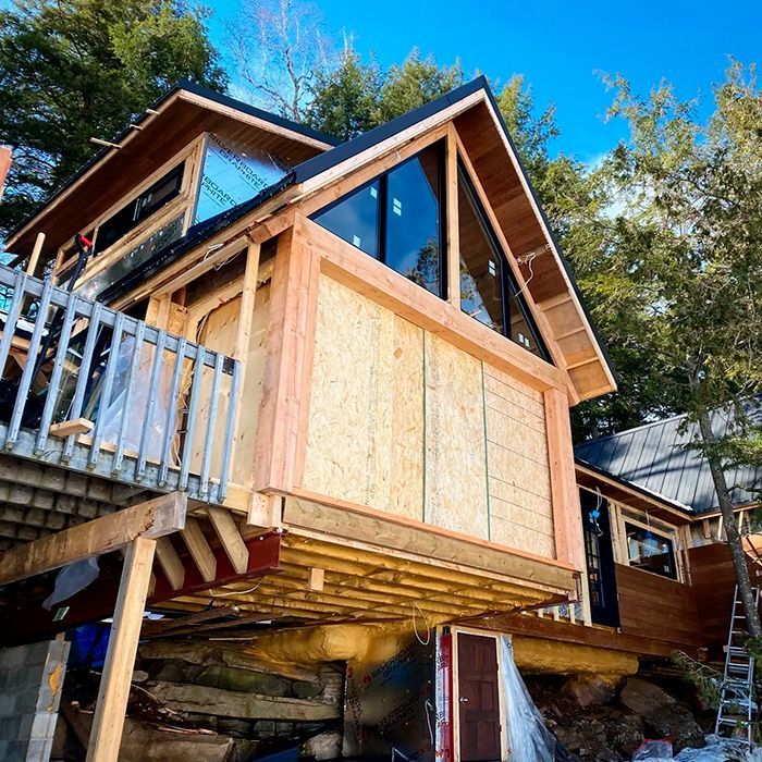 Cabin under construction with wooden framing, large windows, and deck, set on a rocky hillside.