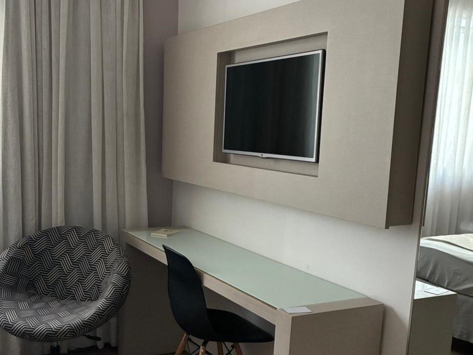 Escrivaninha moderna de quarto de hotel com TV fixada na parede, cadeira preta e poltrona estampada ao lado das cortinas.