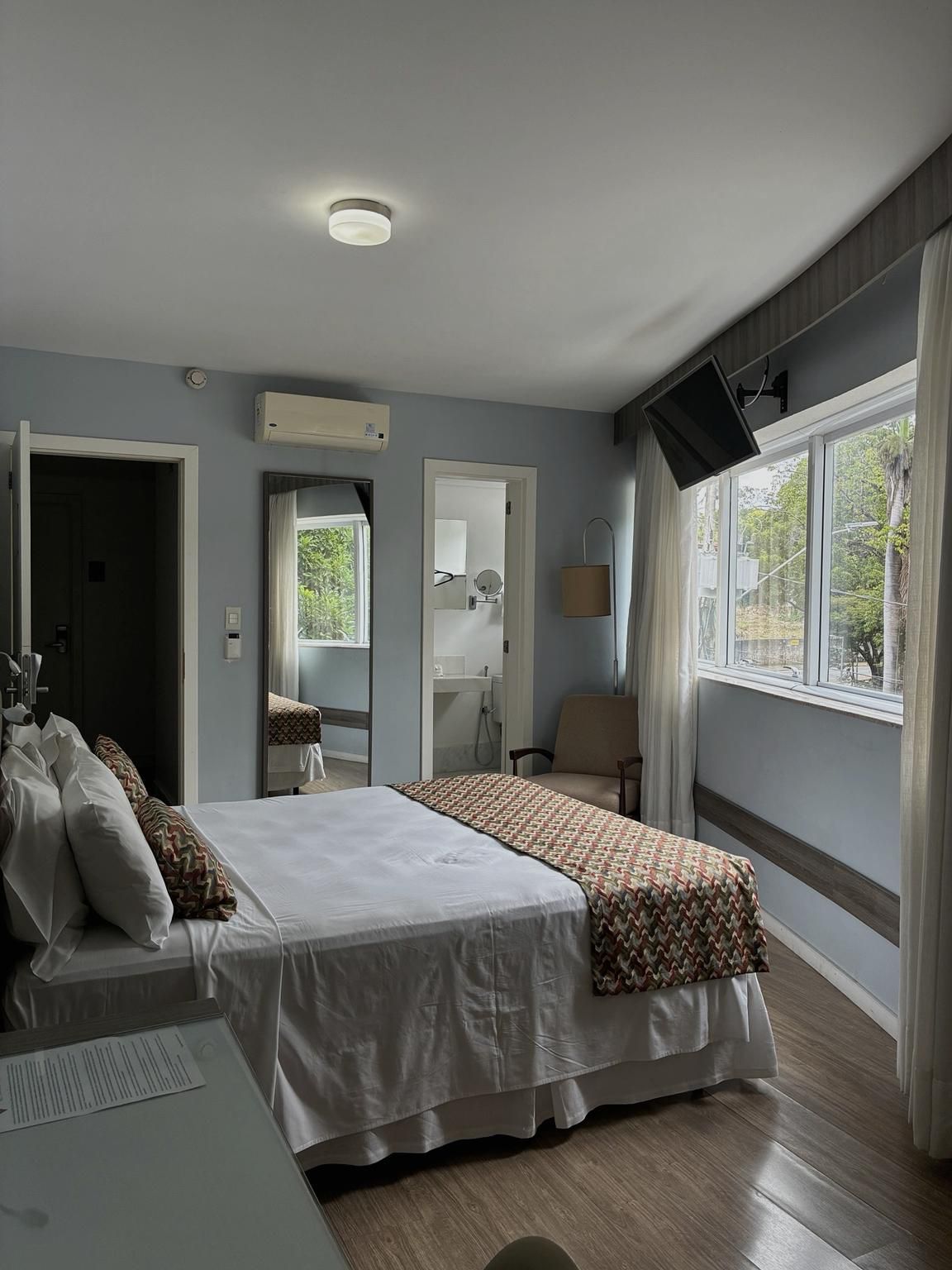 Quarto iluminado com cama, janelas amplas, paredes cinza e ar-condicionado de parede.