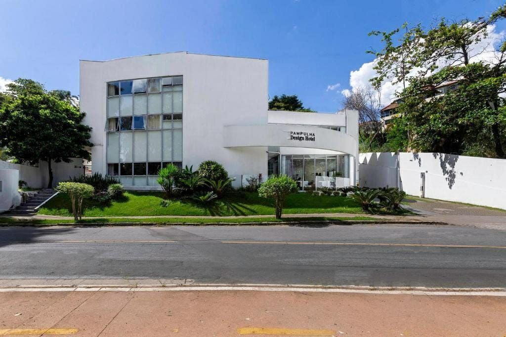 Prédio branco de dois andares com janelas de vidro, uma placa na entrada e árvores ao longo da estrada.