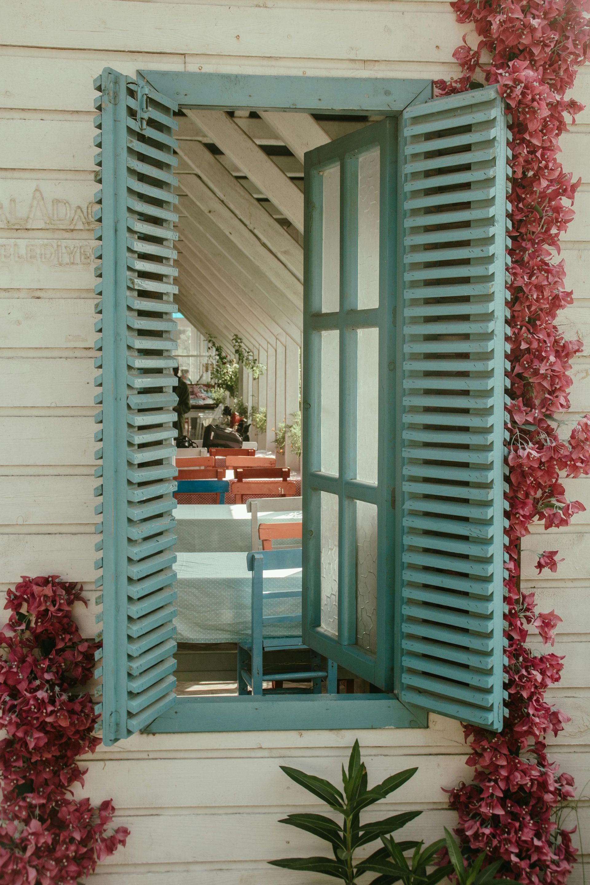 A window with blue shutters on a white wall with pink flowers growing on it.