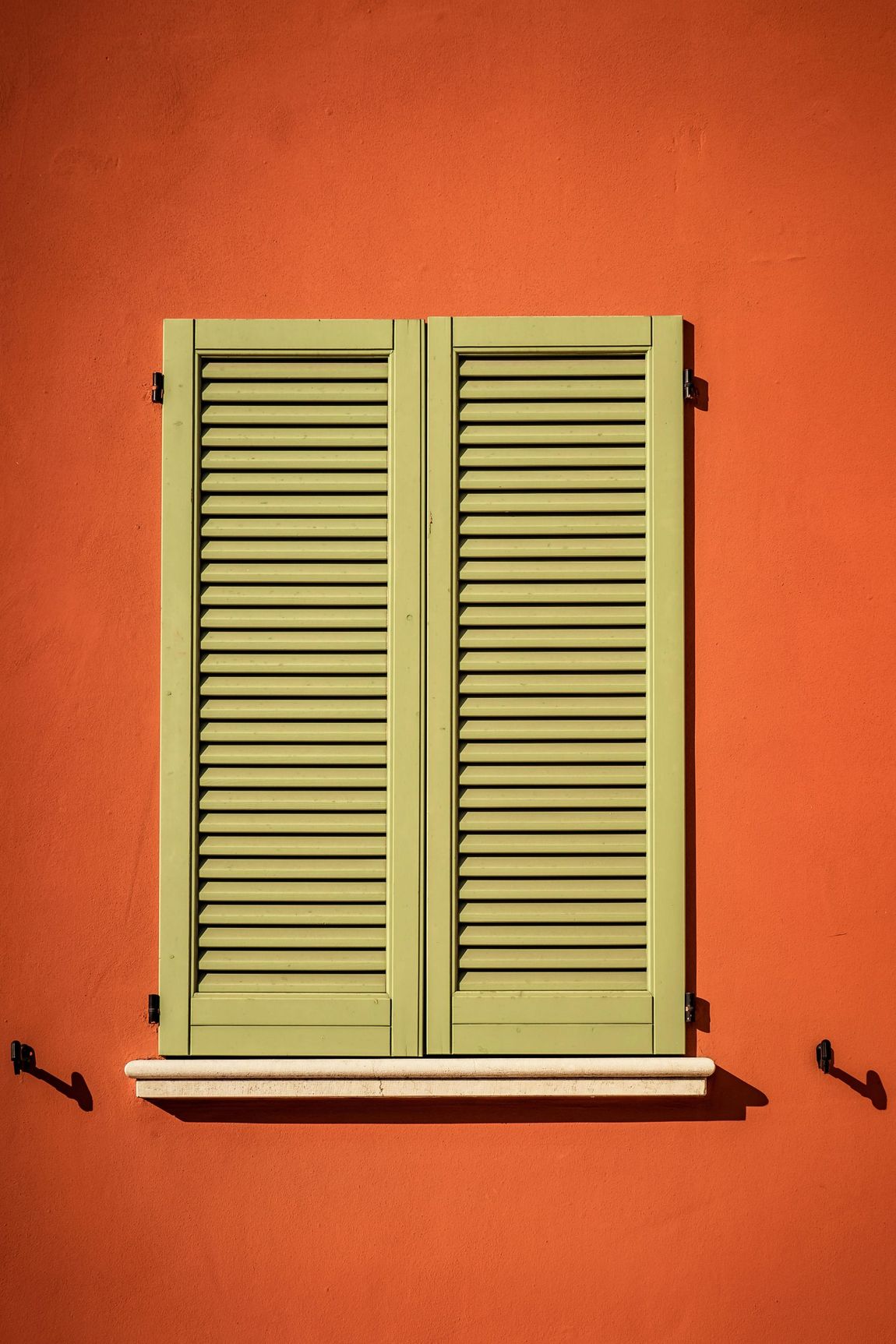 A green window with shutters on an orange wall.