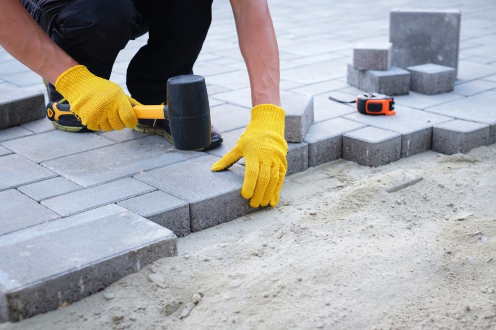 A man wearing yellow gloves is using a rubber mallet on a brick walkway.