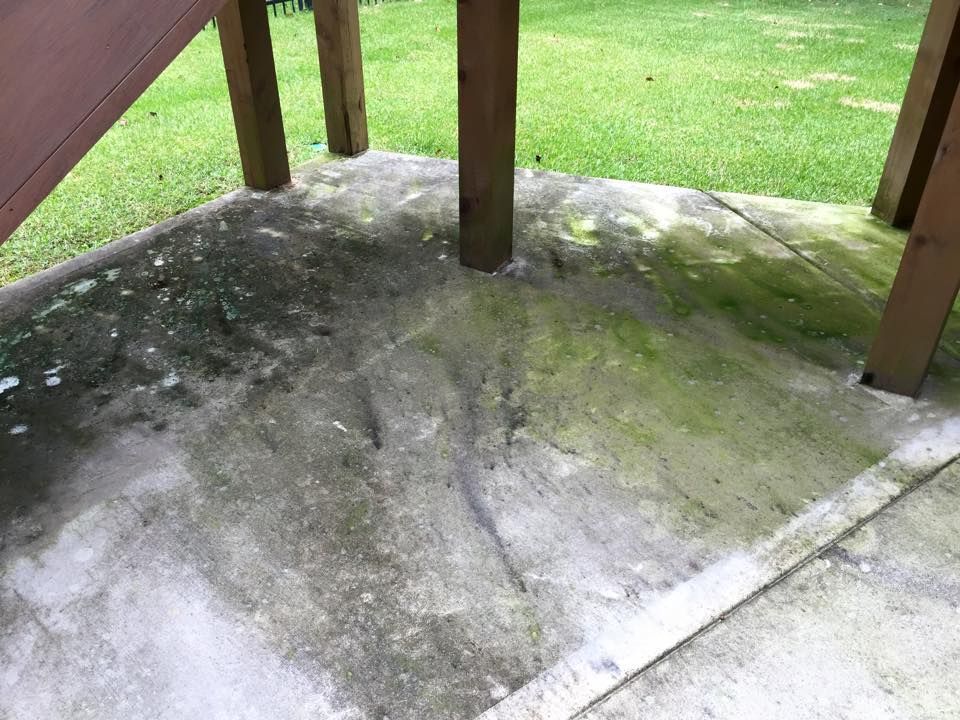 A dirty concrete patio under a wooden deck with moss growing on it.