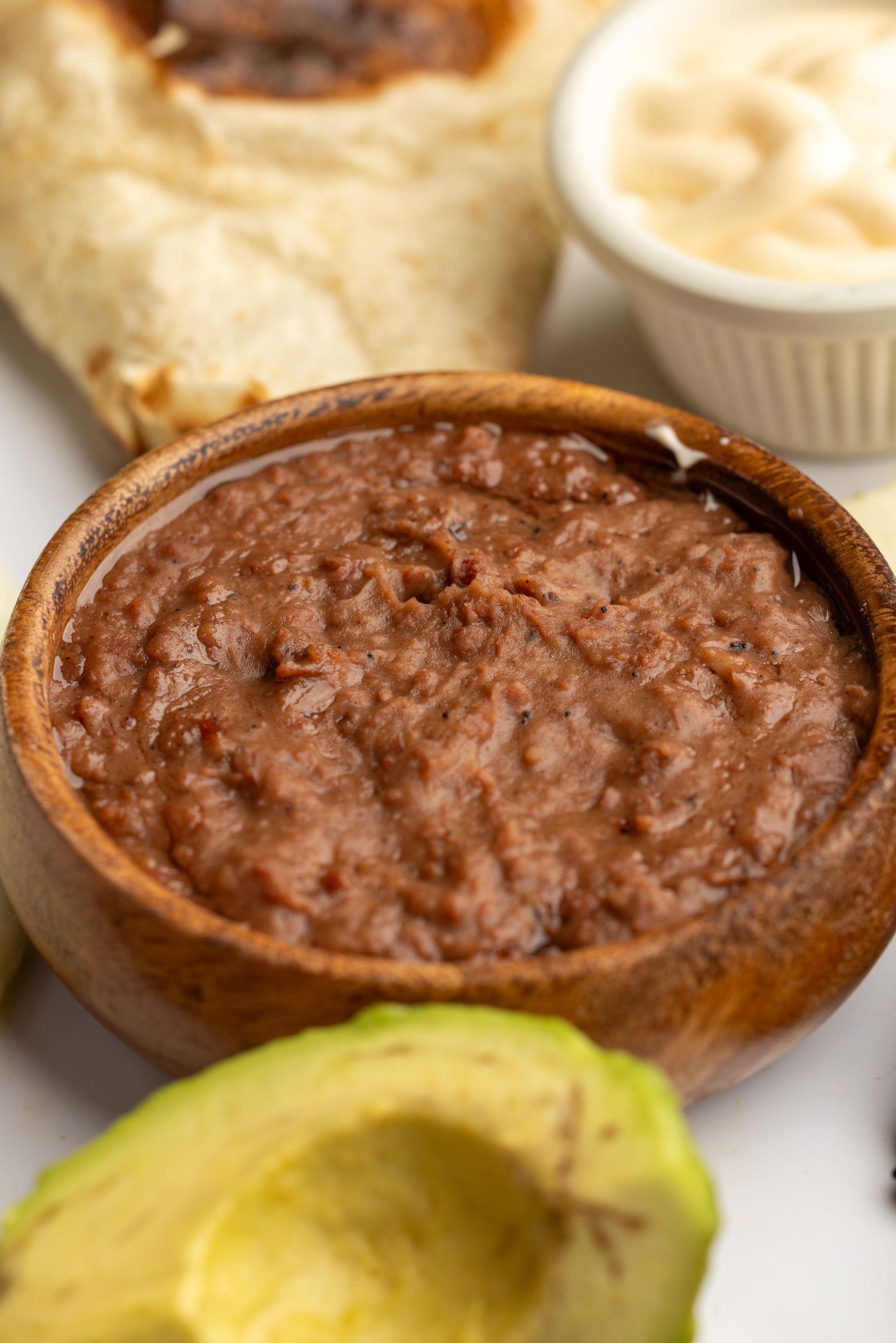 A wooden bowl filled with beans next to an avocado and a tortilla.