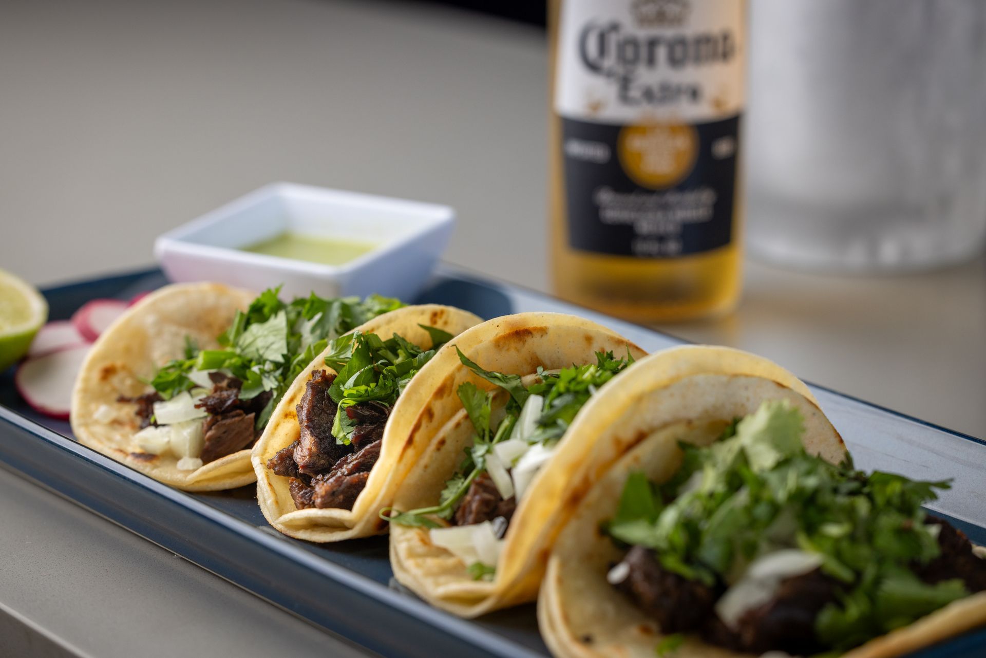 Tacos with cilantro, radishes, and green sauce on a rectangular plate with a Corona beer in the background.