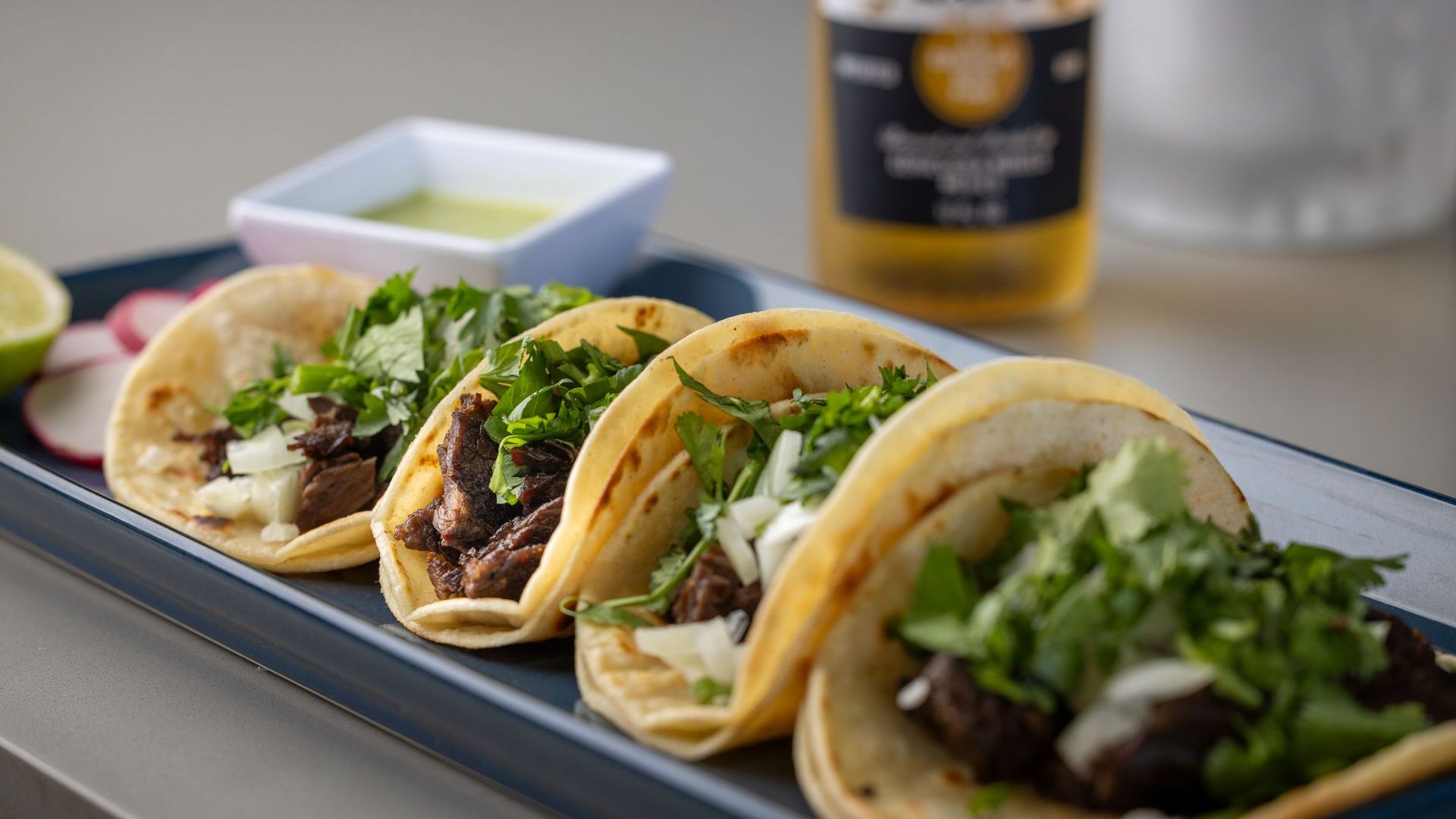 A plate of tacos with a bottle of beer in the background.