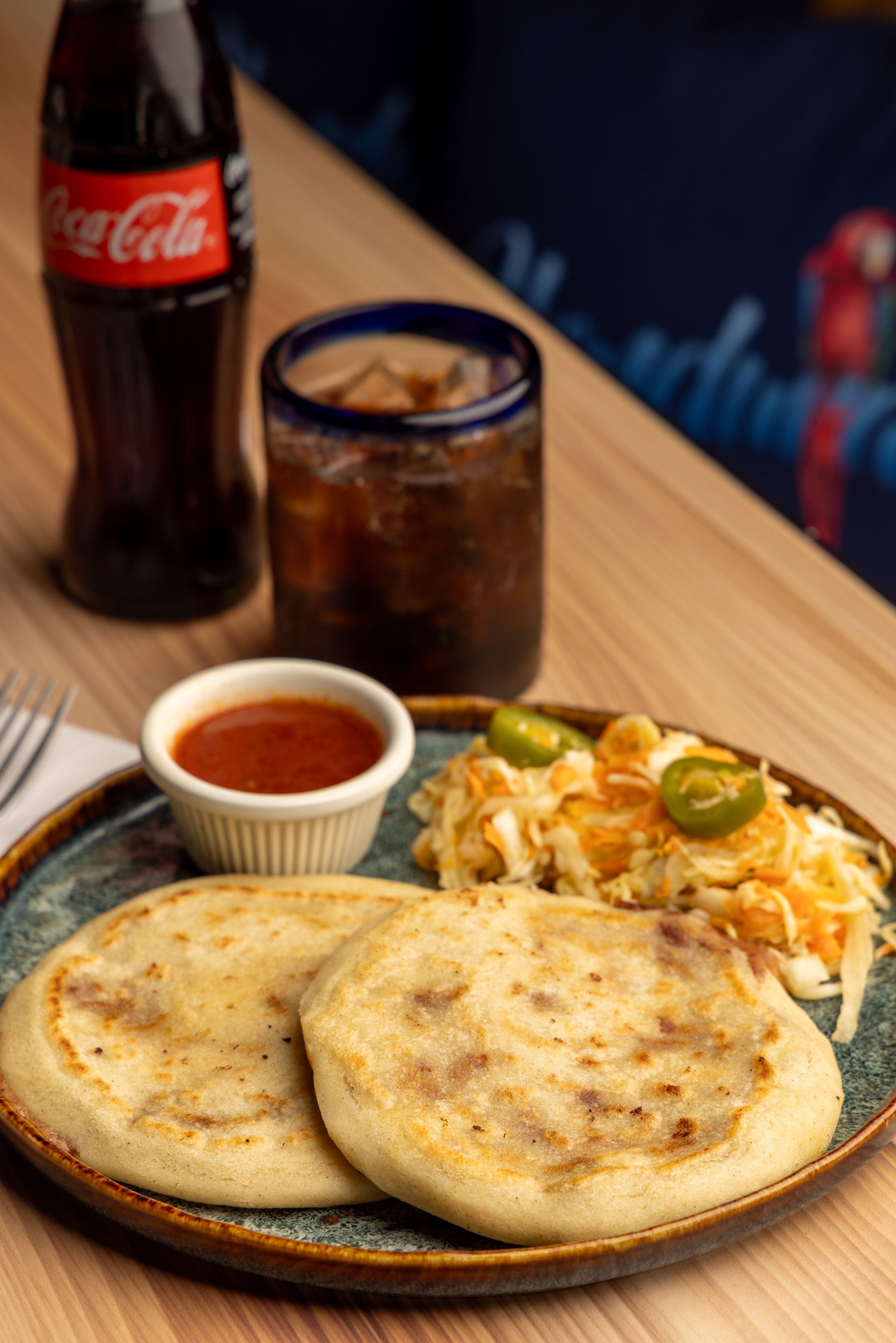 Two pupusas on a plate with coleslaw, sauce, a drink, and a bottle of Coca-Cola.