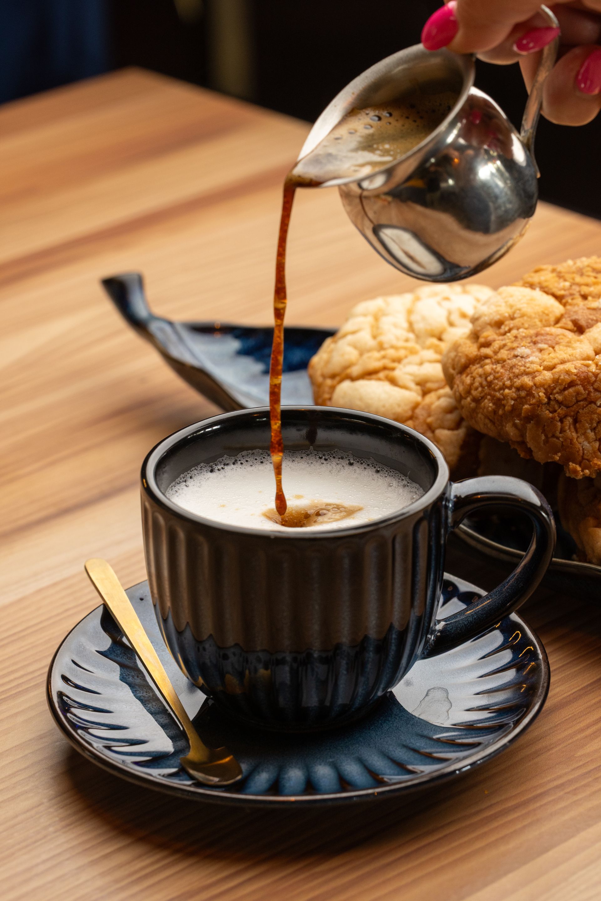 Coffee being poured into a blue-rimmed cup on a saucer. Beside it are pastries, on a wooden table.