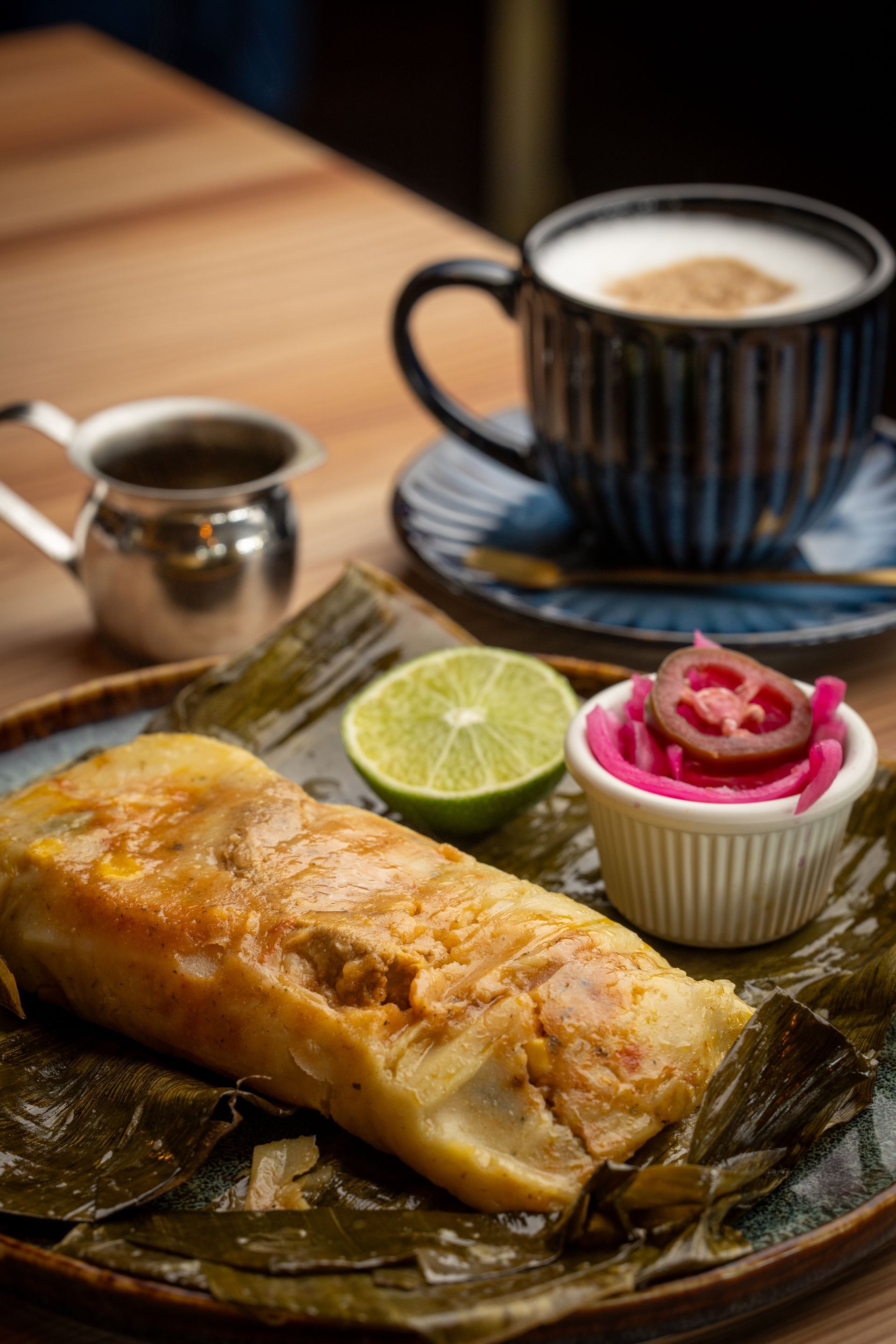 Tamale on a plate with lime, pickled onions, and a cappuccino on a wooden table.