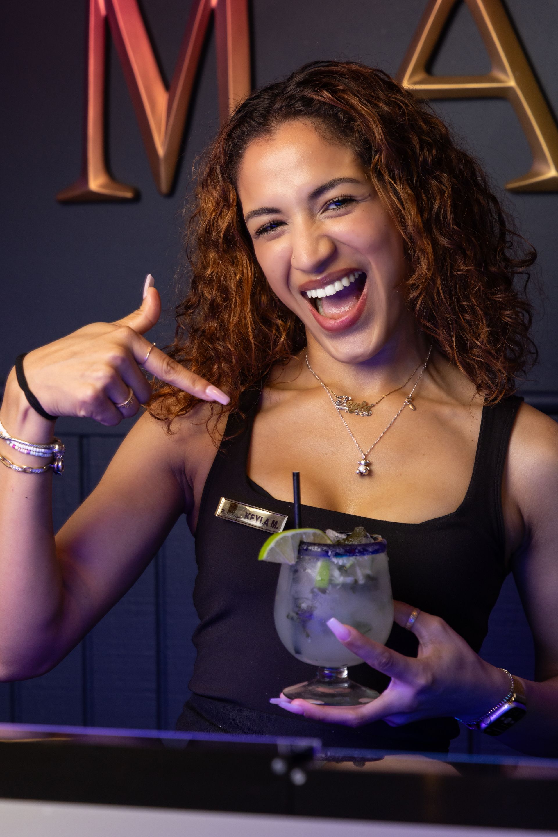 Woman pointing at margarita, smiling. Bar setting with dark background and gold lettering.