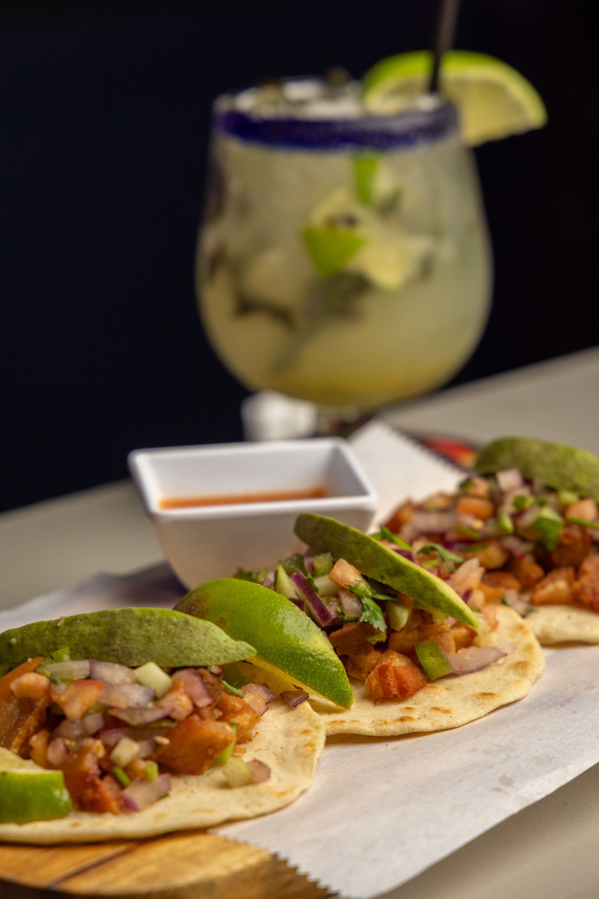 Tacos with salsa, avocado slices, and lime wedges on a wooden board, with a margarita in the background.