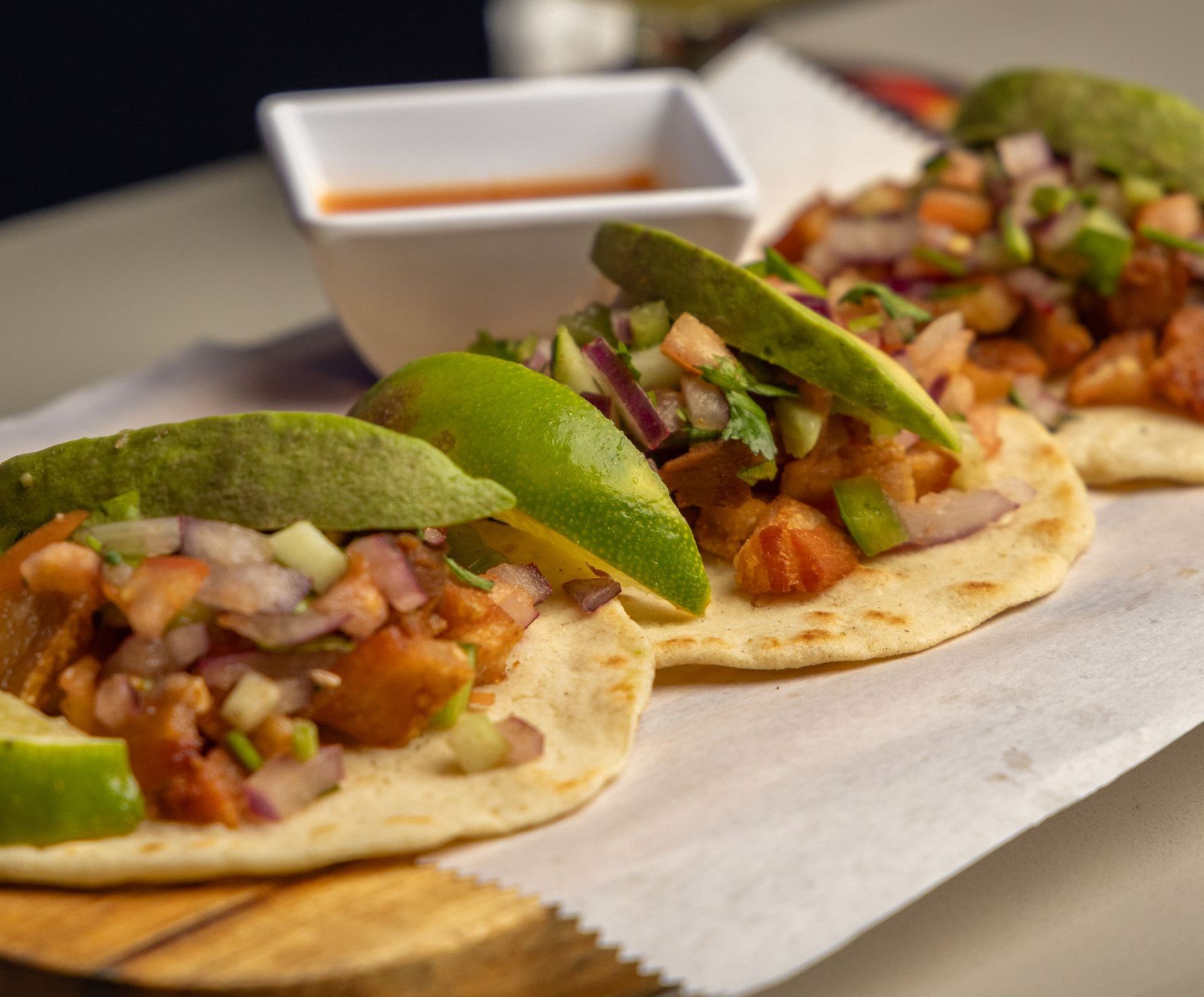 Three tacos on a wooden board with salsa and lime, topped with avocado and pico de gallo.