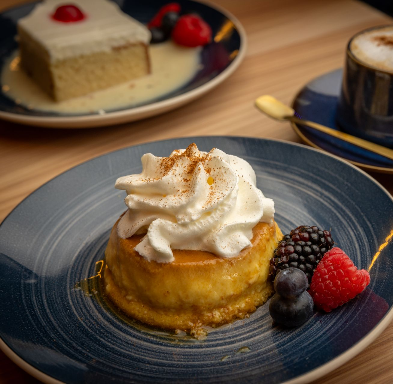 Flan topped with whipped cream and berries on a blue plate, with cake and coffee in the background.