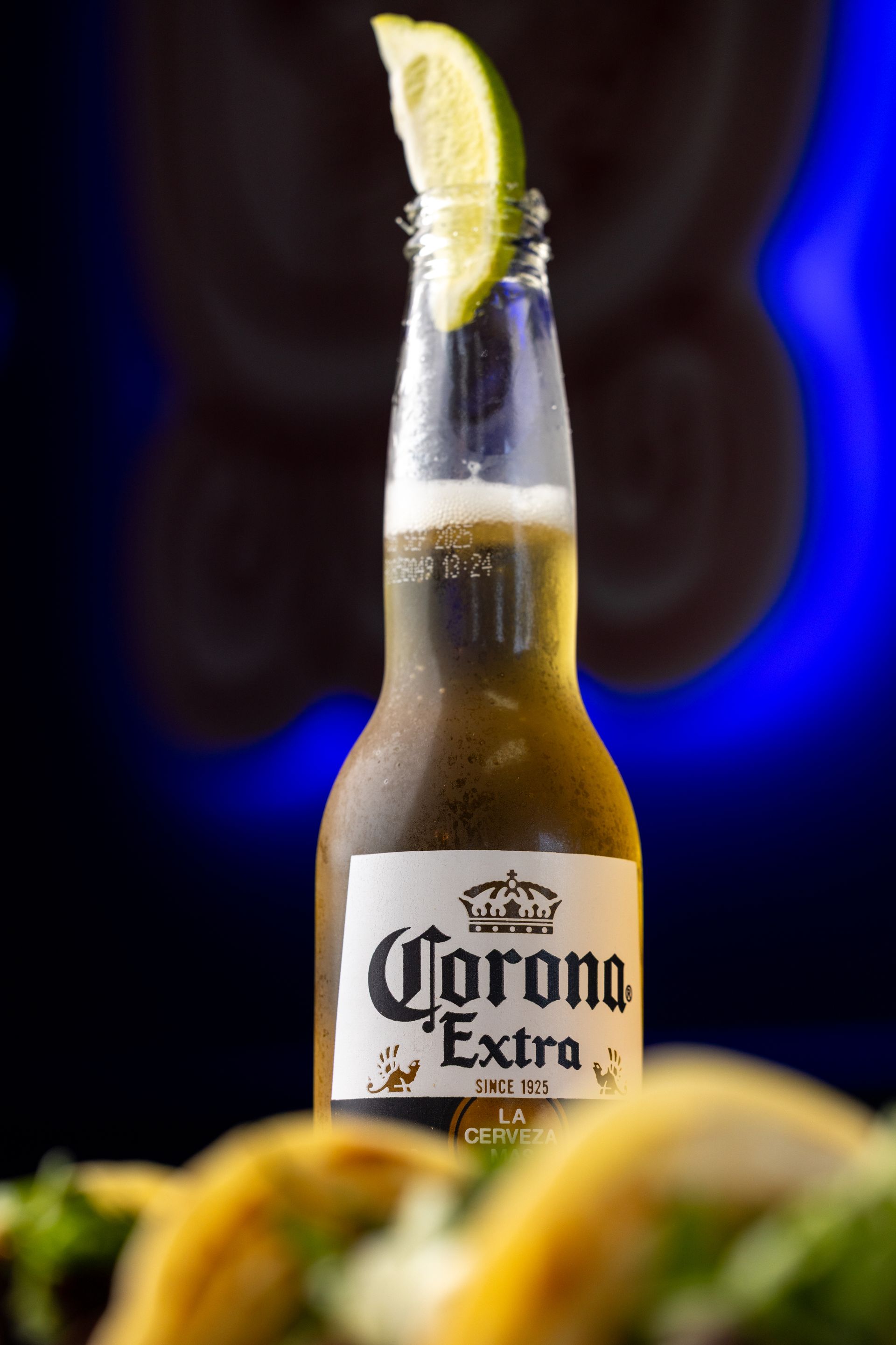 Corona Extra beer bottle with lime wedge, tacos in front, blue background.