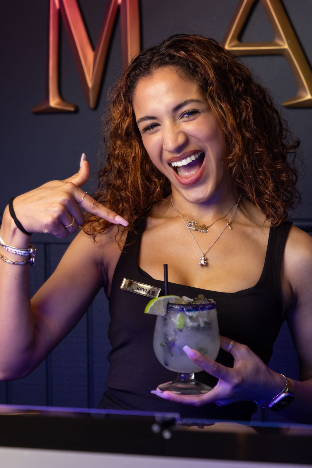 Woman pointing at a margarita, smiling, with curly brown hair, in a bar setting.