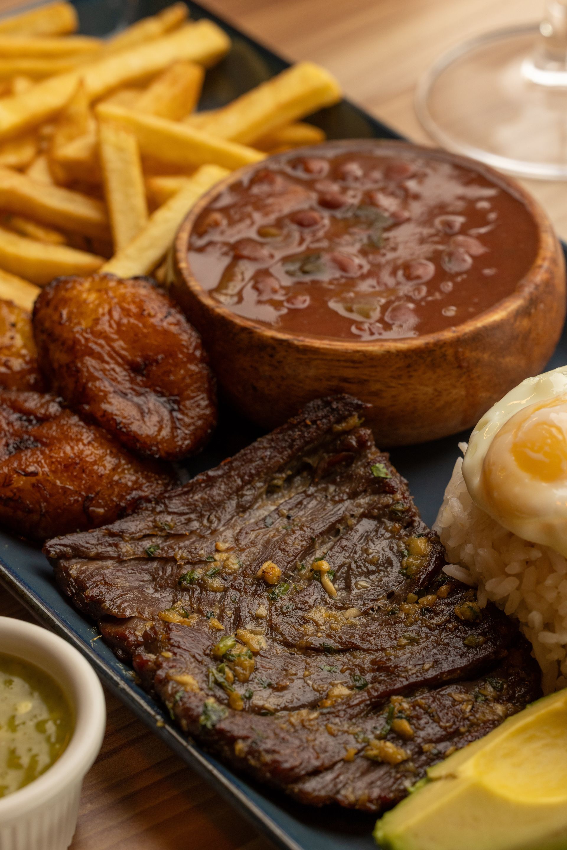 Grilled steak with beans, fried plantains, fries, egg, rice, avocado, and sauce on a plate.