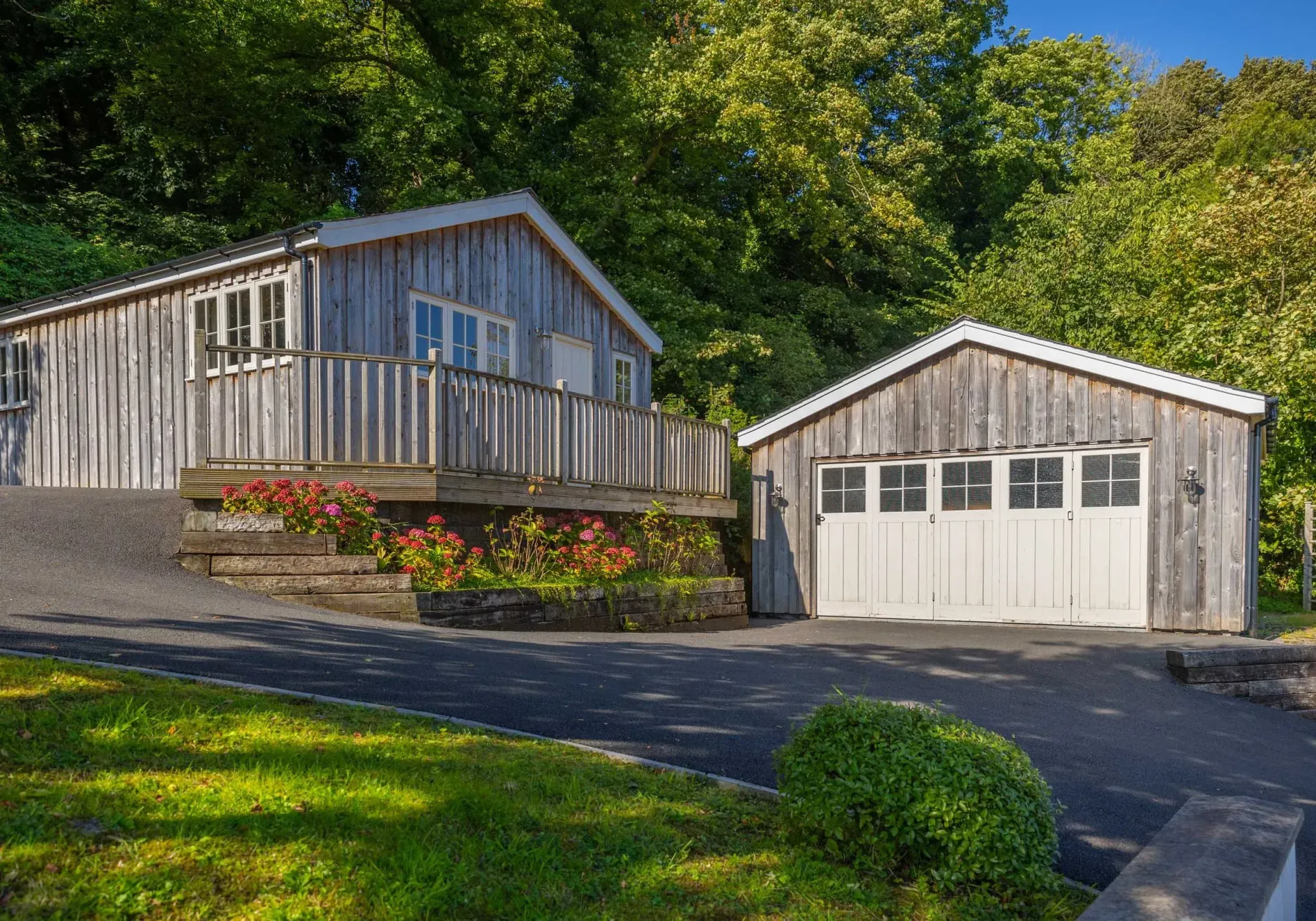 Wooden cabin and garage with a driveway, surrounded by green foliage, under a blue sky.
